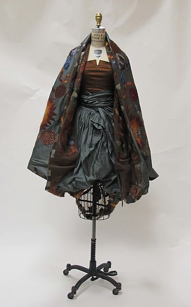 a dress made with a patchwork of colorful, floral fabric, layered over a grey, draped skirt, is displayed on a rolling mannequin against a white background. the dress is styled in a vintage style, with a loose fit and a flowing, asymmetrical silhouette. it has a unique and interesting design, with a variety of different fabrics and patterns being used.
