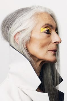 a mature woman with silver hair and bright yellow eyeshadow looks into the camera. she is wearing a white coat with a black lining and has grey hair that is styled straight and long.