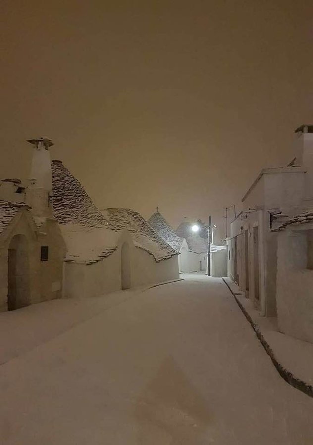 the image shows a deserted street in alberobello, italy, covered in snow. the iconic trulli houses with their distinctive coned roofs are also blanketed in snow, creating a winter scene at night. a street lamp in the distance illuminates the narrow street, lined with the whitewashed trulli buildings. the houses feature chimneys and unique architectural details.