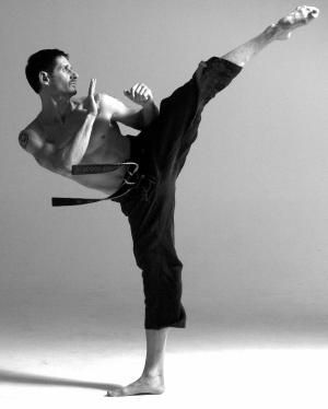 a man in a black karate gi is midkick, with his leg extended high and his body contorted in a powerful pose. he's wearing a black belt, and his expression is focused and determined. the background is a simple white studio backdrop, highlighting the athlete's form and movement. the image is in black and white, creating a timeless and classic feel.  the overall image conveys a sense of strength, discipline, and athleticism.