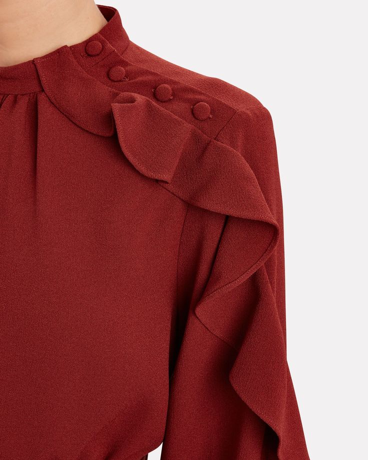 the image is a closeup shot of a woman's maroon dress. the dress features a high neck, long sleeves with ruffles cascading down the shoulders and arms, and subtle buttons along the neckline.  the fabric appears to be a textured, possibly woven material with a solid reddishbrown color. the overall style is elegant and sophisticated.  only a portion of the dress and a person's neck and shoulder are visible. the background is a plain offwhite, which keeps the focus on the dress's details.