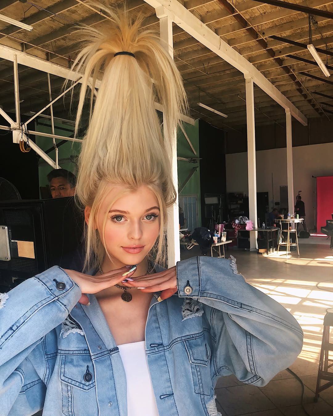 the image shows loren gray in a studio setting. she has blonde hair styled in a tall ponytail, wears a light blue denim jacket over a white top, and accessorizes with a necklace. she is posing with her hands near her face and looking at the camera. the background reveals a typical production set with lighting equipment and crew members present.