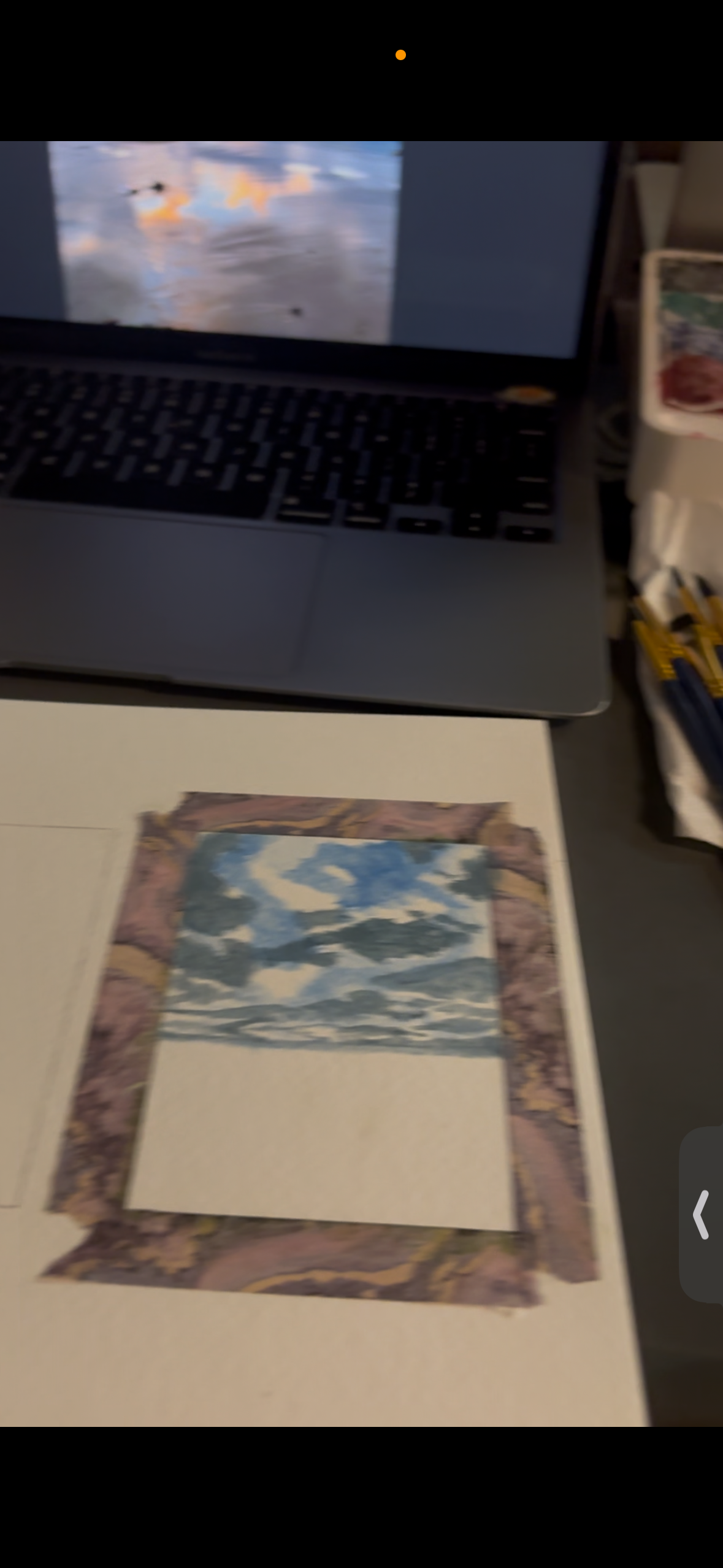 the image shows a desk with a laptop, a watercolor painting taped to a larger piece of paper, and some art supplies. the painting appears to depict a sea scene with clouds and sky. the laptop screen displays a similar image, possibly the reference for the painting.