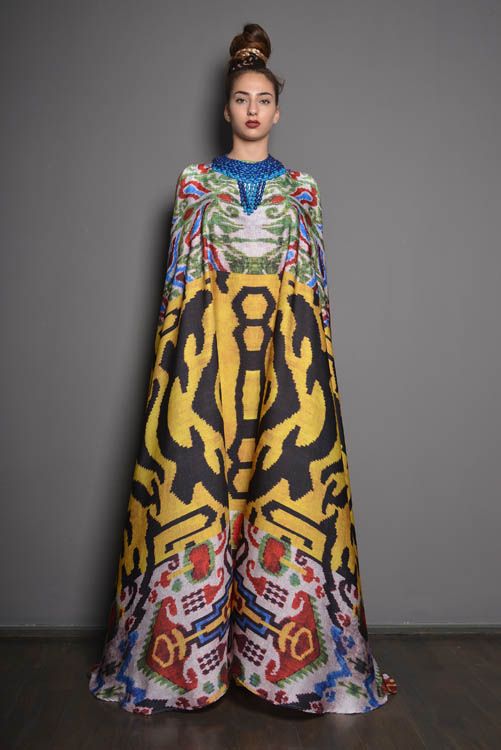 a fulllength studio shot of a female model wearing a striking maxi dress. the dress is a vibrant mix of colors and patterns, featuring a dominant yellow and black ikatinspired design, accented by red, blue, and green geometric patterns.  the neckline includes a beaded embellishment. the model is positioned against a plain gray background, her hair styled in a high bun. the overall impression is one of high fashion and elegance.