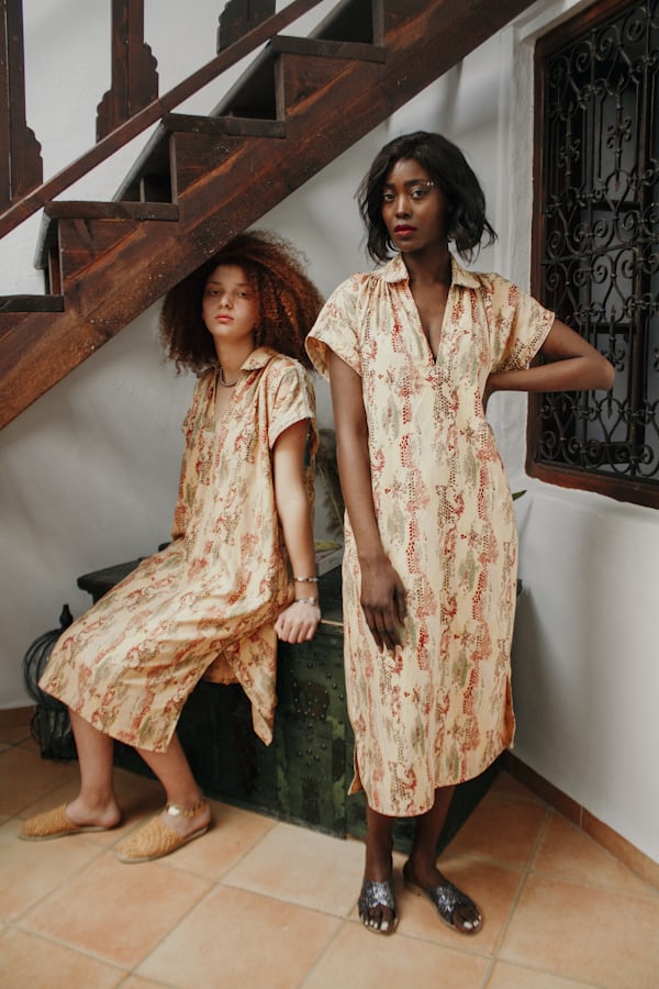 two women in matching patterned dresses are standing by a wooden staircase in a room with a moroccanstyle door.  they are both smiling and looking at the camera.  one of the women is sitting on a dark green chest, and both models are wearing sandals. the dresses are a light beige color with a repeating pattern in oranges and reds. the overall aesthetic is casual, yet elegant and slightly bohemian.