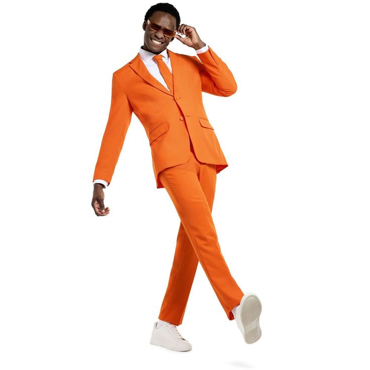 the image shows a stylish man in a bright orange slimfit suit. he's smiling and playfully adjusting his sunglasses. he's wearing white sneakers and is captured midstep, giving a dynamic and energetic feel to the shot. the background is plain white, allowing the bold orange suit to be the clear focal point.  the overall impression is one of modern, confident, and vibrant style.