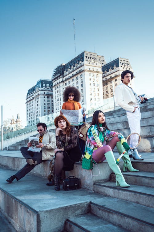 a diverse group of stylish young adults, dressed in 70sinspired retro clothing, are sitting on a set of stairs in an urban setting.  the background features impressive city buildings, possibly in buenos aires, argentina. they appear relaxed and confident, with some holding maps and a tennis racket, suggesting a sense of travel and leisure.  their attire showcases individualistic fashion choices reflecting a vintage 70s aesthetic.