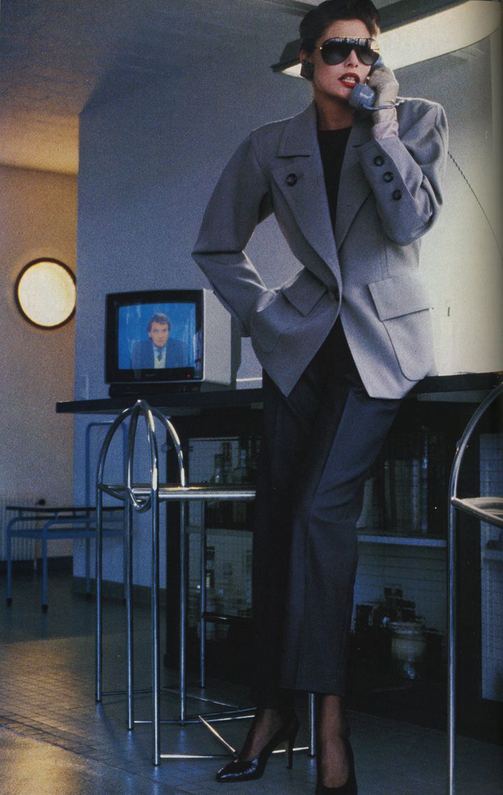 a stylish woman in a gray pantsuit and sunglasses is on the phone in a modern apartment.  she's standing next to a chrome bar cart and a television showing a news broadcast. the setting is minimalist and the overall aesthetic is very 1980s. the woman exudes confidence and sophistication.