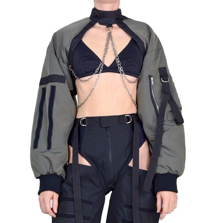 a woman is wearing a green bomber jacket with black accents, a black bralette, black pants with cutouts, a silver chain choker, and a silver body chain. she is posing in a studio with a white background. the jacket has a unique design with a high collar and long sleeves. the pants have a low rise and are fitted through the legs. the chain choker is simple and delicate, while the body chain is more elaborate and eyecatching. the woman's overall look is edgy and stylish.