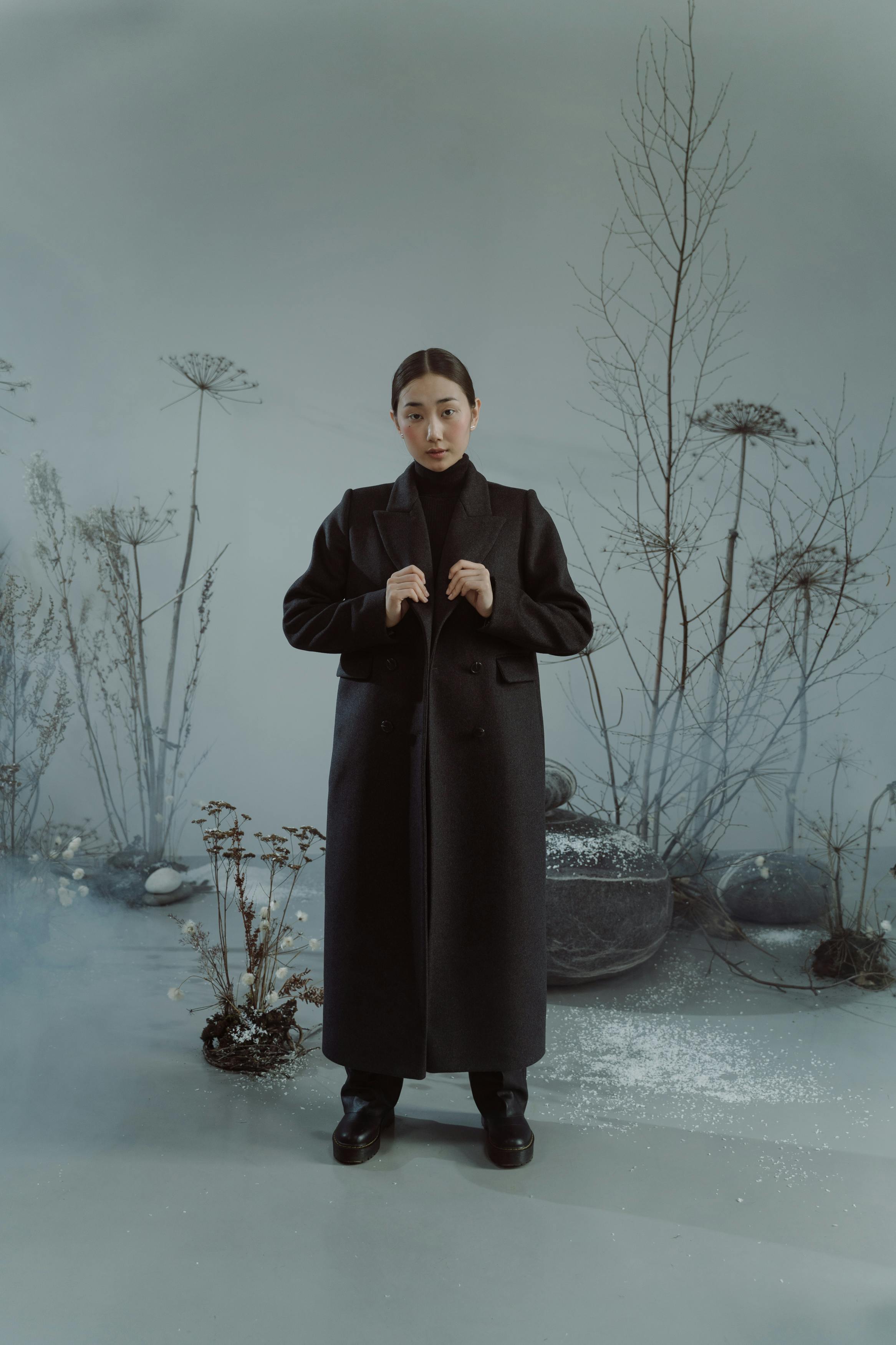 the image shows a fulllength portrait of a young asian woman, standing in a long black overcoat. she has a serious expression on her face and her hands are clasped in front of her. she is standing in a desolate winter scene, with dry plants and a foggy atmosphere creating a moody and atmospheric feel. the background is muted in tones of gray and blue, providing a minimalist contrast to the woman's black coat. the overall aesthetic is elegant and stylish.