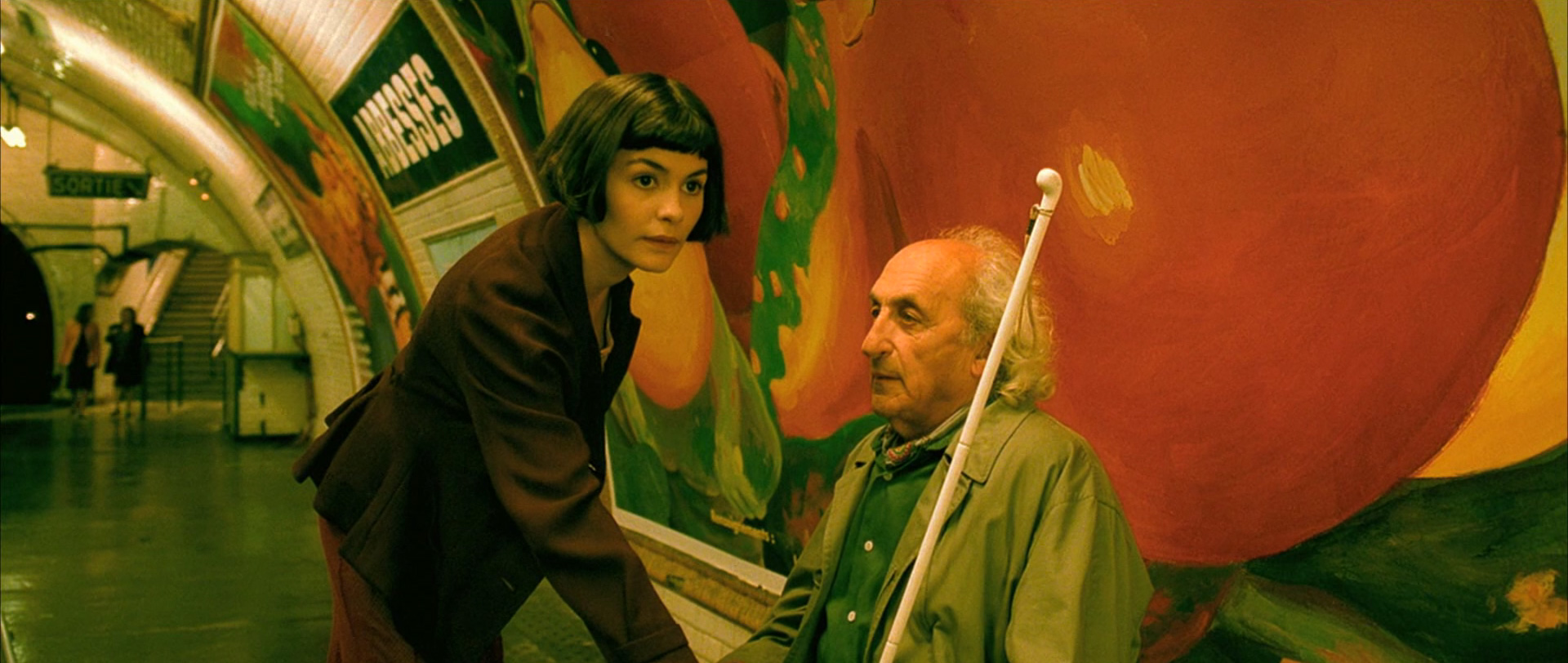 a young woman with short, dark hair and a maroon jacket leans towards an elderly man with a white cane in a brightly lit subway station. they appear to be engaged in conversation. the wall behind them features a vibrant mural with abstract shapes in orange, yellow, and red hues.