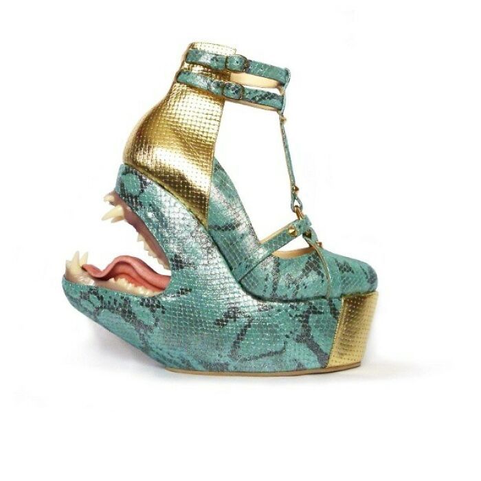 a single, highheeled shoe with a monstrous design is displayed against a white background. the shoe features a platform base and a wedge heel, both adorned with a gold finish. the main body of the shoe is patterned with blue and dark grey snake skin print. the toe area is designed to resemble the head of a monster, complete with sharp, menacing teeth and a pink tongue. the shoe also has a golden ankle strap and tstrap for fastening.