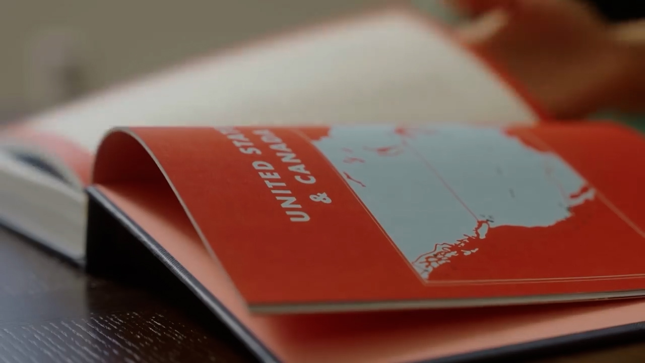 a closeup shot of an open atlas to the united states and canada section. the atlas is red and has a map of north america on the right page. the atlas is lying on a dark brown table. the image is brightly lit with natural light, suggesting it was taken indoors during the day.