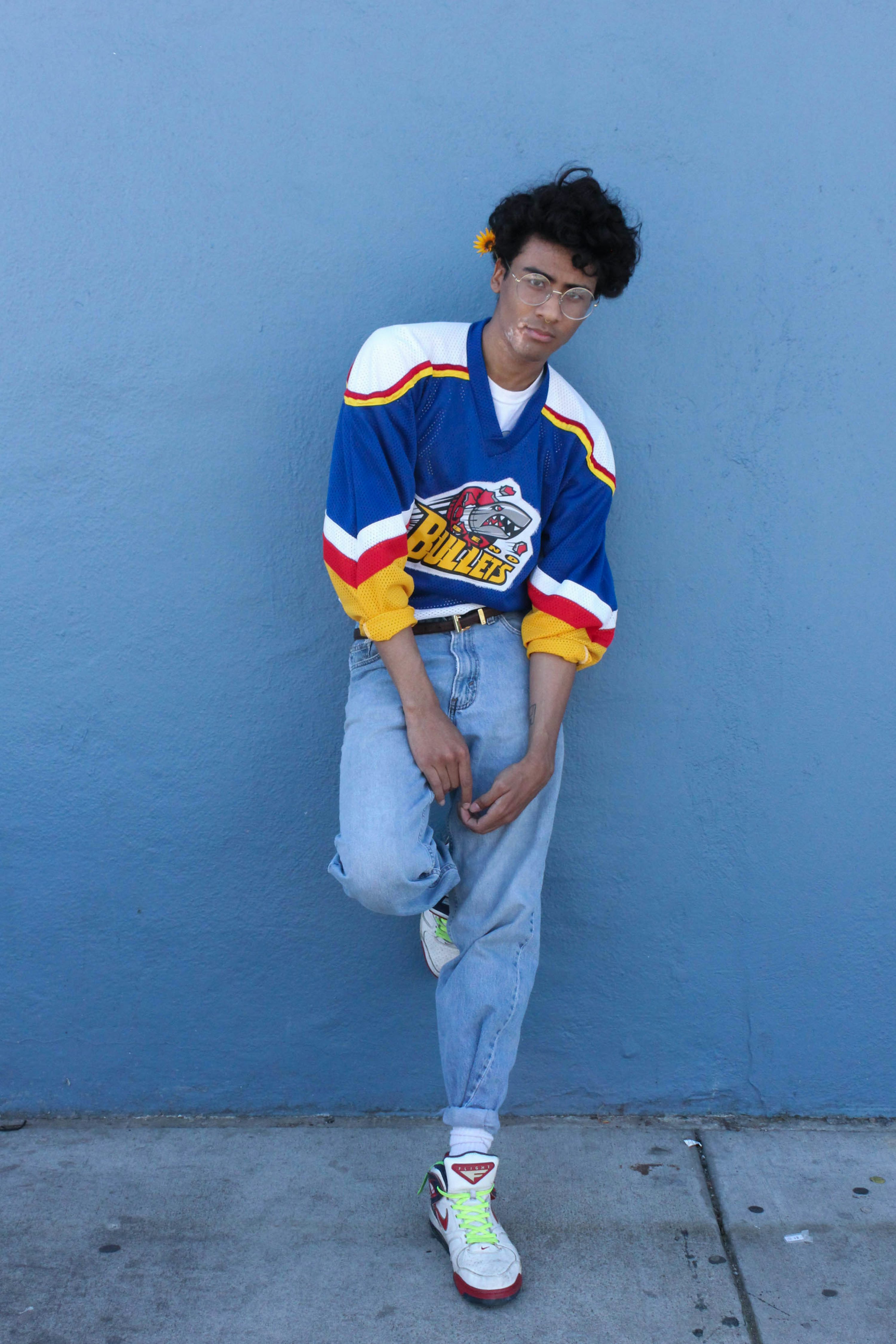 a young man stands casually against a blue wall. he's wearing a vintagestyle blue hockey jersey with red and yellow stripes, light blue baggy jeans cuffed at the ankles, white hightop sneakers with neon green laces, and glasses. he has curly brown hair. the overall style is reminiscent of 90s fashion.  he appears relaxed and confident.