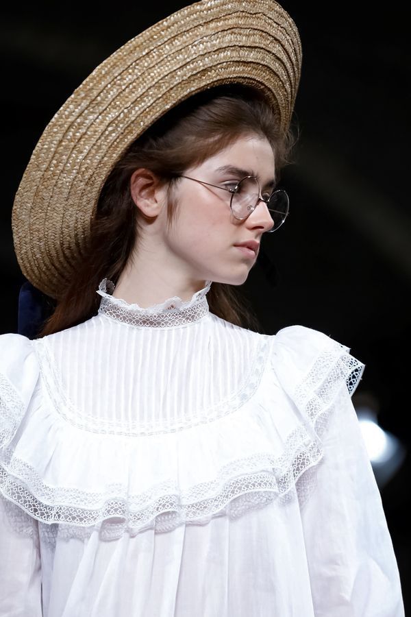 closeup shot of a fashion model wearing a white dress with lace details and ruffles. the dress has a high neck and pleated bodice. the model also wears a large straw hat and round glasses. the overall style is vintage or retro, elegant and sophisticated. the background is dark and out of focus, drawing attention to the model and her attire. the image likely comes from a fashion show or photoshoot.