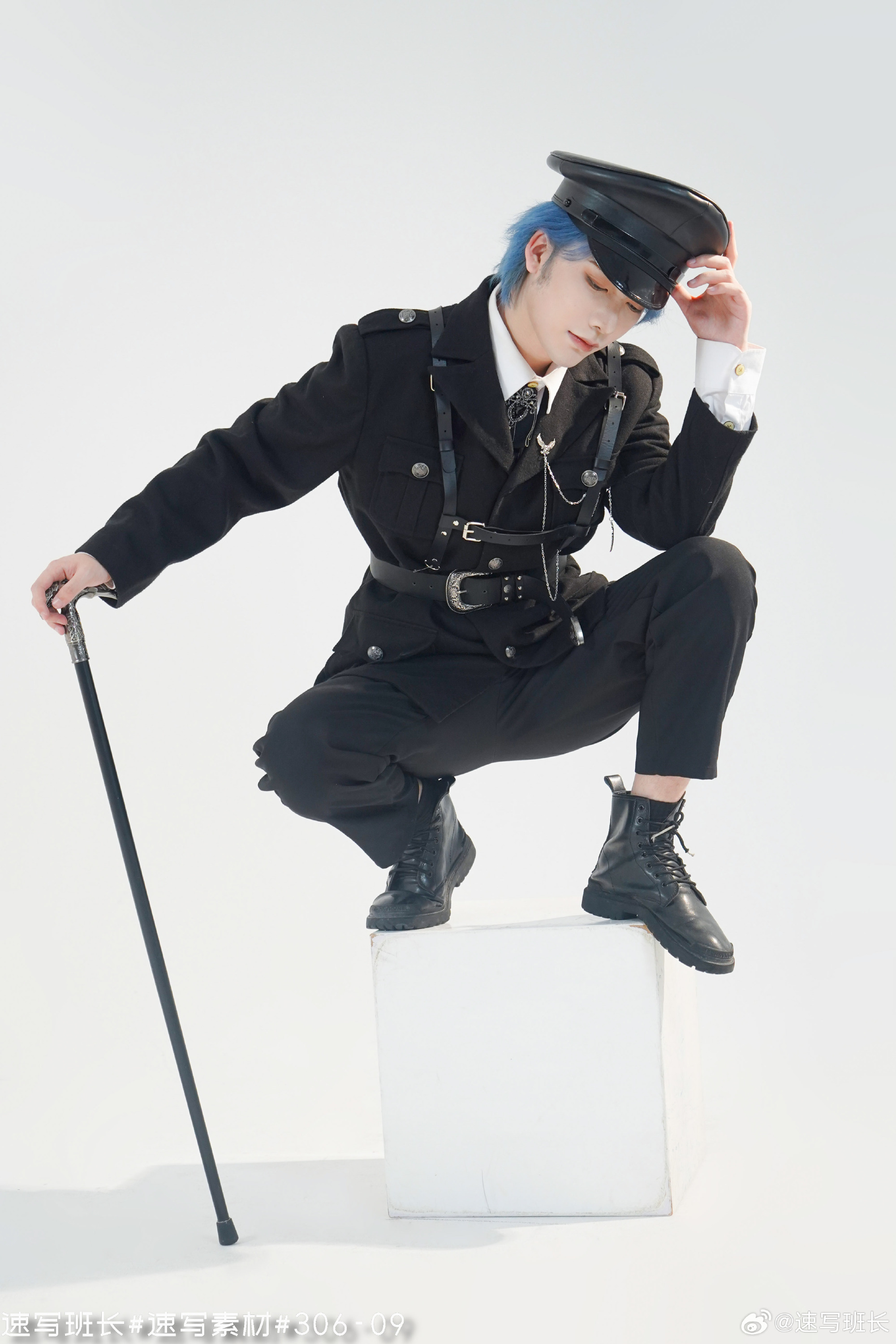a young man with blue hair, wearing a black militaryinspired uniform with silver accents, poses on a white cube against a white background in a studio setting. he holds a black cane in his right hand and touches his cap with his left.  the uniform includes a jacket with epaulets and decorative belts, black pants, a white shirt, a black tie, and black boots. his expression is serious and stylish.