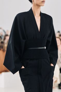a woman in a black long sleeve dress with a black belt. the dress is styled with a deep vneck and a cinched waist. the woman is posing in a chic and sophisticated manner.
