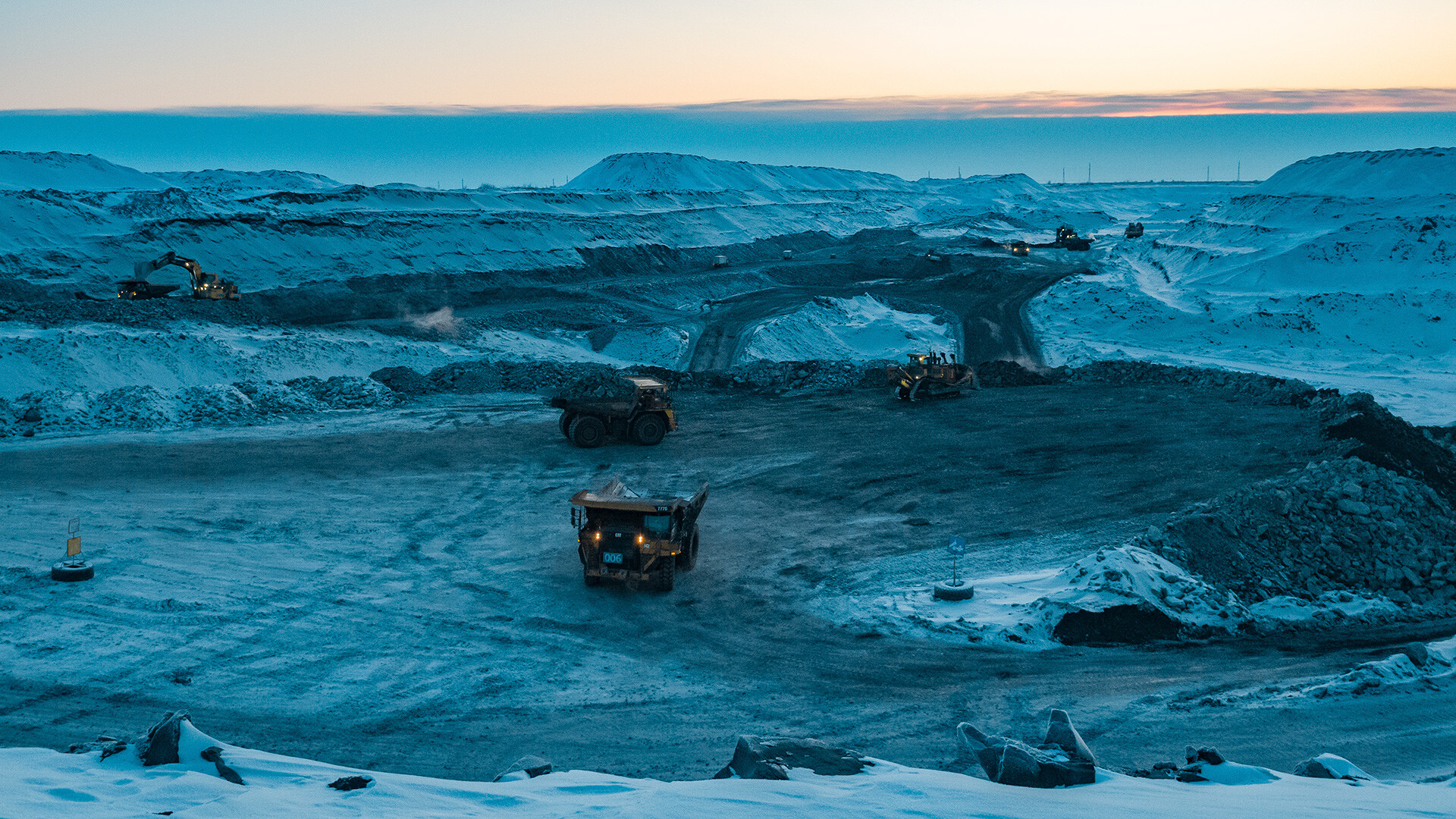 an aerial view of a mining operation in a snowcovered open pit quarry. heavy equipment, including excavators and dump trucks, are working in the quarry. the sky is a light blue color, and the snowcovered hills in the background are illuminated by the setting sun. the overall tone of the image is cold and industrial.