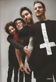 a group of four young men, possibly a band, pose together in a photo. they are all wearing dark clothing and one man has a tattoo on his arm and is wearing a shirt with a white symbol on it. they are all looking at the camera and seem to be having fun.