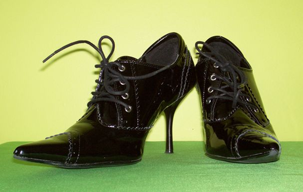 two black, patent leather, laceup high heels are displayed on a green surface against a light green background. the shoes have pointed toes and stiletto heels. they are laced with black laces, and the shine of the patent leather is visible.