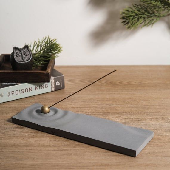 the image shows a stylish, gray concrete incense burner with a rippled surface. a single incense stick rests on the burner, which features a small brass ball at the point where the incense rests. the burner sits on a lightbrown wooden table next to a book titled "the poison king", a small wooden tray with a black owl figurine, and a pine branch. the overall scene is minimalist, serene and suggests a calming atmosphere.