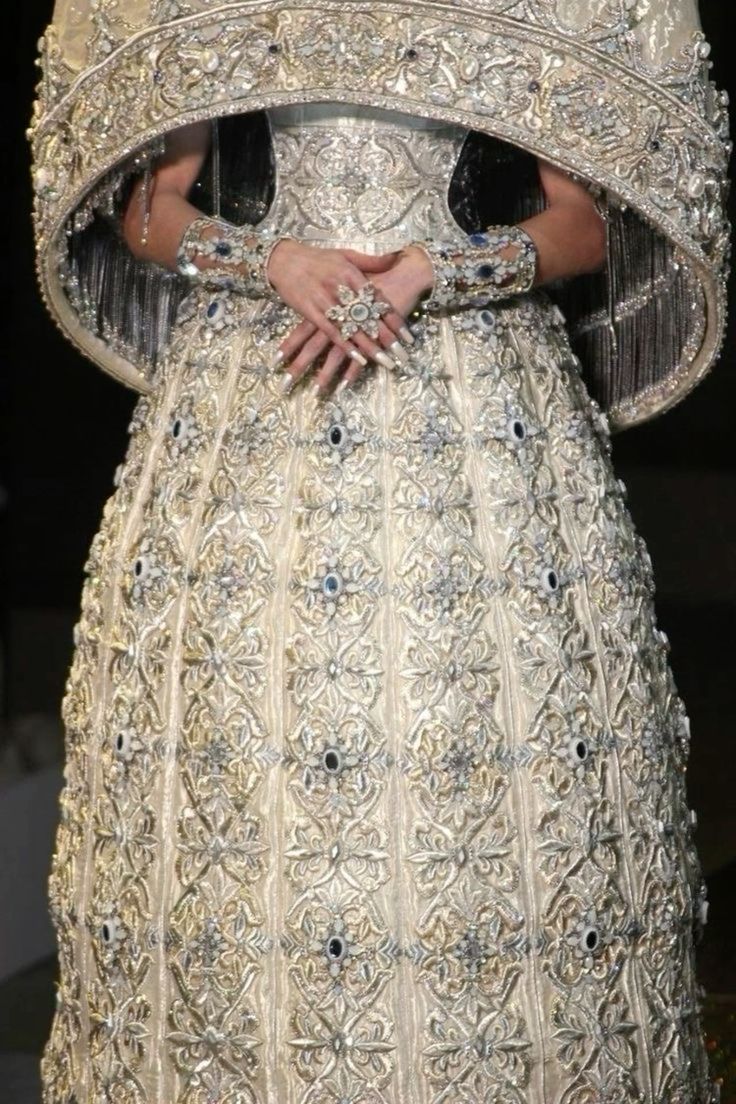 a closeup shot of a model wearing an extravagant white gown. the dress is adorned with intricate gold and silver embroidery, creating a luxurious and glamorous look. the model's hands are clasped together, showcasing the ornate jewelry on her fingers. the dress's design and detailing suggest it's a highfashion piece, likely from a runway show or a prestigious designer.