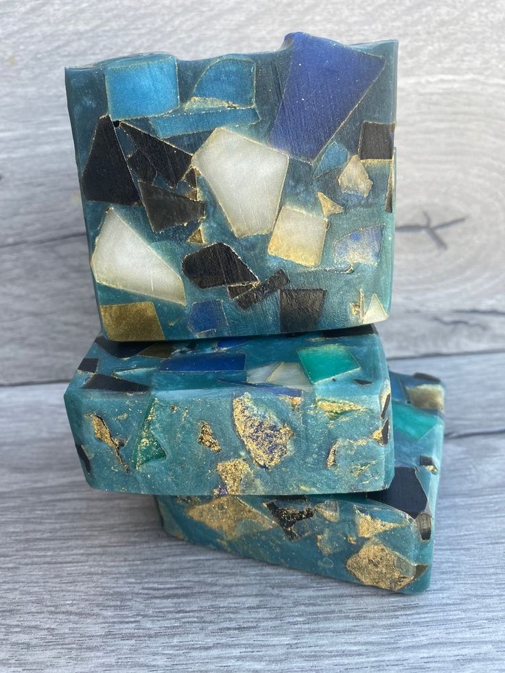 a stack of three handmade soap bars with a mosaic design featuring blue, green, and gold accents. the soap bars are made with a unique and beautiful mosaic design, with small pieces of colored soap creating a textured and artistic surface. the soap bars are neatly stacked on top of each other, showcasing their intricate design and inviting you to experience their luxurious scent and gentle lather.