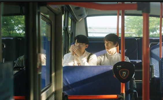 two young men sit on a bus, lost in their own thoughts. they are bathed in warm sunlight streaming through the windows, creating a peaceful and contemplative atmosphere.