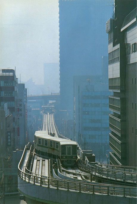 a vintage photograph of a tokyo monorail train navigating a curved elevated track amidst a dense cityscape of towering skyscrapers. the image is slightly hazy or foggy, lending a nostalgic and atmospheric quality. the train is lightcolored, contrasting with the darker buildings.  the photograph suggests a scene from the 1970s or 1980s, showcasing the rapid urbanization of tokyo and the integration of advanced public transportation systems. the perspective is from a high vantage point, looking down on the train and the surrounding urban environment.