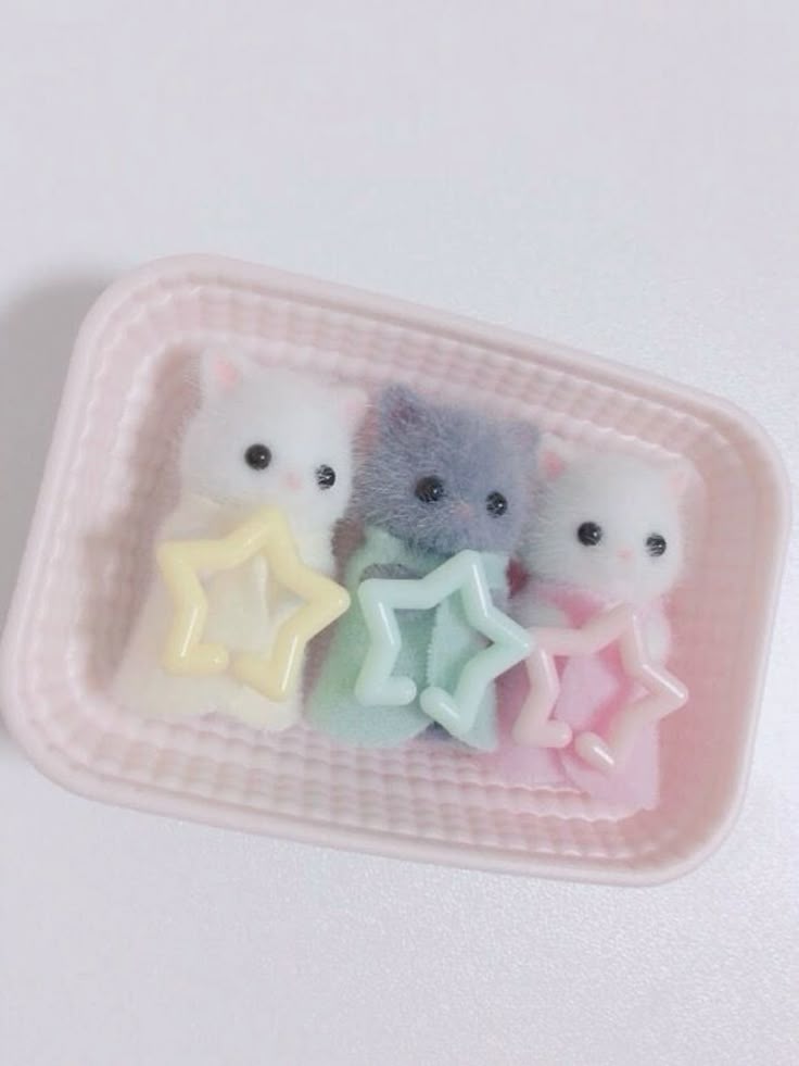 three miniature cat figurines in a pink, woven plastic basket. each cat is a different color—white, gray, and pale pink—and they are each holding a pastelcolored, starshaped toy. the overall aesthetic is cute and delicate.