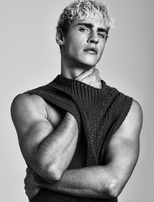 a young, blond man in a studio portrait. he is wearing a knit sweater with a thoughtful expression.  his arms are crossed and he appears to be looking off to the side.  the background is a plain gray.