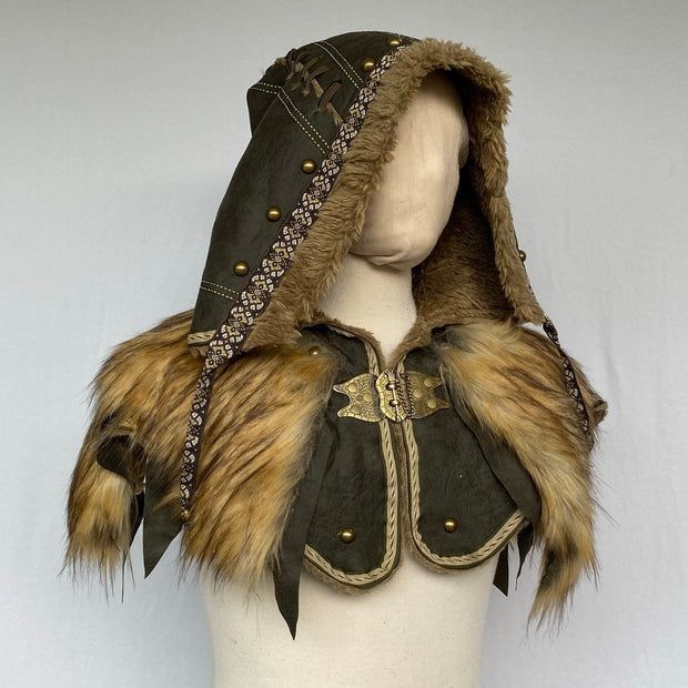 a hooded cape is displayed on a mannequin. the hood is dark green with brass studs and brown trim. the cape is trimmed with tan fur. a brass clasp holds the cape together at the front.