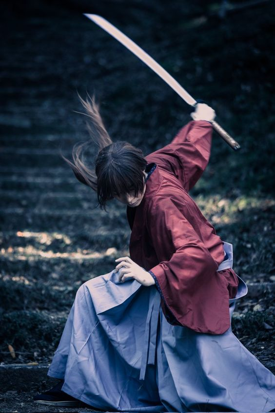 a woman dressed as a samurai in a fighting stance is captured midmotion, dramatically swinging a katana.  she is outdoors, seemingly on worn stone steps with her long dark hair whipping around her head as she moves.