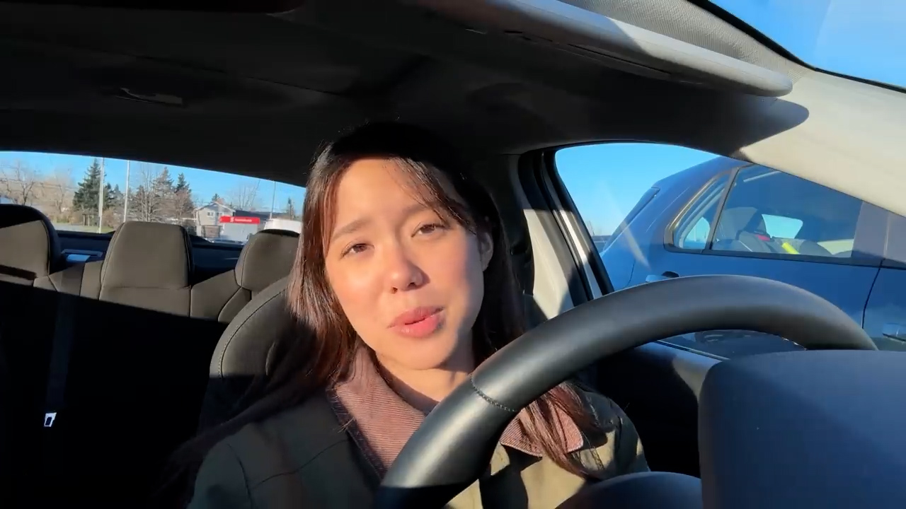 a young asian woman with long dark hair is sitting in the driver's seat of a car and talking to the camera. she is wearing a brown jacket and has a neutral expression on her face. the car is parked in a parking lot and the sun is shining brightly, casting a warm glow on the woman's face.  the image is taken from a firstperson perspective, as if the viewer is sitting in the passenger seat.