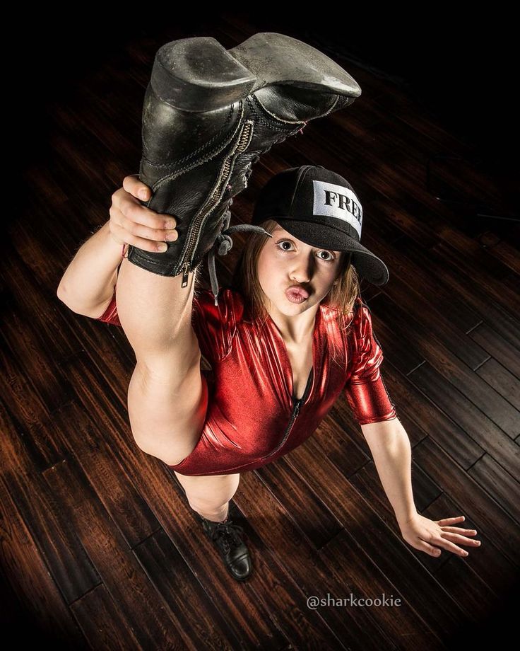 a young woman in a red leotard and black boots is doing a split while holding her foot and making a funny face at the camera. the image is taken from a low angle, making her appear larger than life. she is wearing a black baseball cap with the word "frei" on it. the background is a wooden floor.