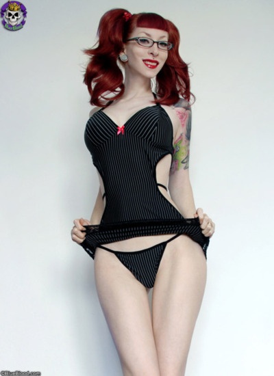 a pinup style woman with red hair and glasses poses in black pinstripe lingerie. she has tattoos on her arm and is wearing red lipstick. her skin is pale. she's standing against a white background.
