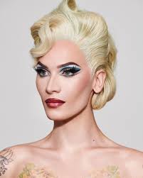 the image is a closeup portrait of a blonde woman with a meticulously styled vintage hairstyle reminiscent of the 1940s or 1950s. her makeup is dramatic, featuring winged eyeliner and a bold lipstick. she has pale skin and subtle tattoos are visible on her chest and shoulders. the background is plain light grey, drawing focus to the model and her striking features. the overall aesthetic is glamorous and elegant.