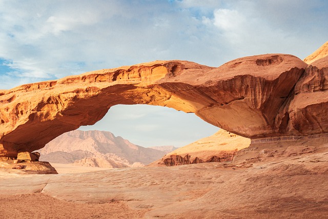 a large natural rock arch dominates the center of the image. the arch is a reddishbrown color and has a smooth, curved underside. the arch is set against a backdrop of a desert landscape, with mountains in the distance. the sky is blue with some white clouds. the lighting is bright and sunny, casting shadows from the arch onto the desert floor.