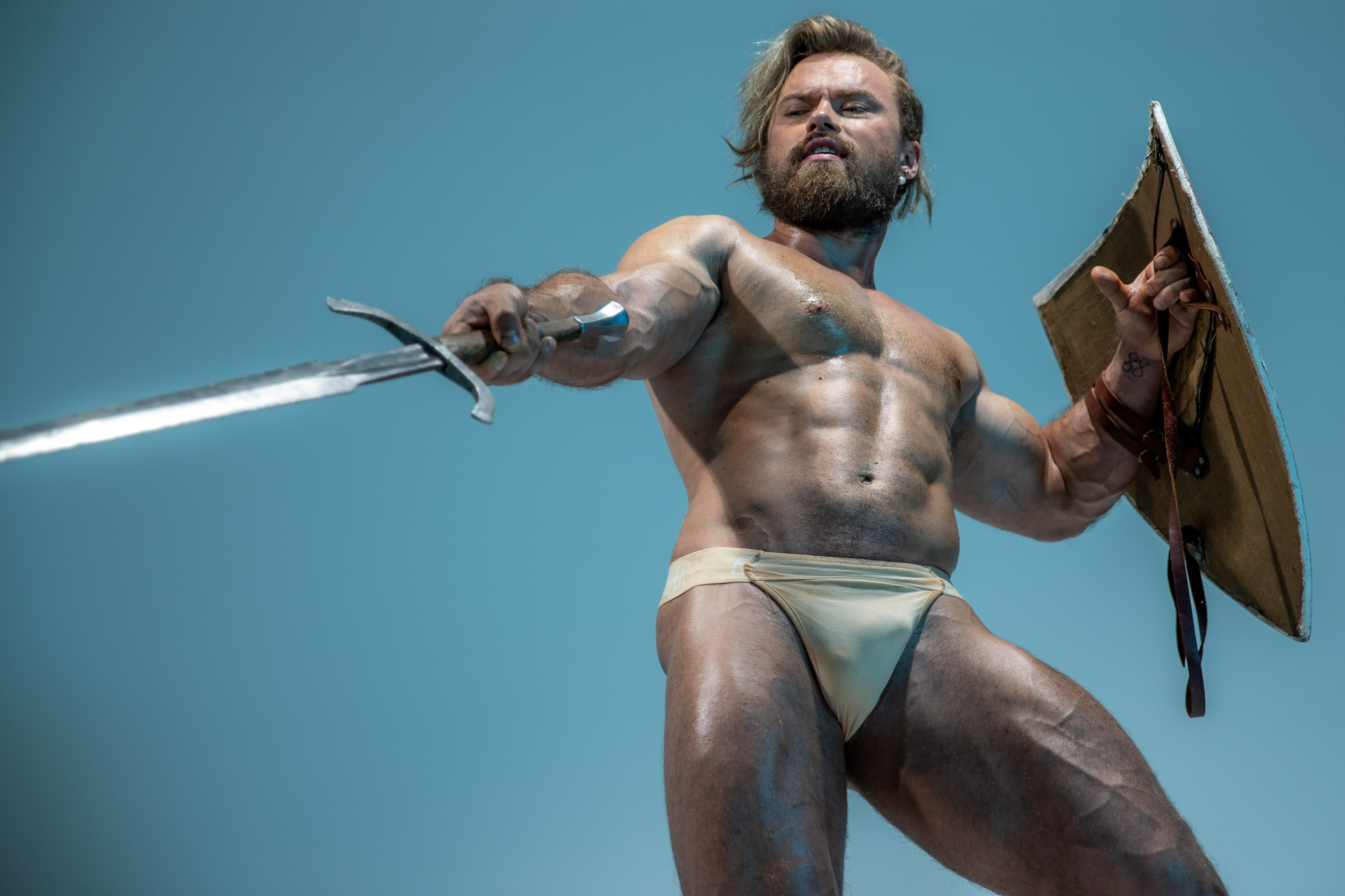 a muscular, barechested warrior stands holding a gleaming sword in one hand and a round shield in the other. he is illuminated by the bright sun against a clear blue sky. the perspective is from below, emphasizing his powerful physique and creating a sense of imposing strength.  he appears ready for battle.