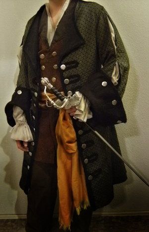 a person dressed in a pirate costume stands facing forward, holding a sword. the costume includes a patterned green and black jacket with large buttons, a brown vest with smaller buttons, a white frilly shirt, and a brown scarf. the sword is silver and black, and the person is also wearing brown pants.