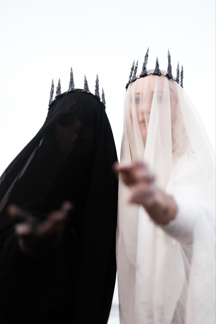 two women stand side by side, one in an all black dress and veil, the other in a white dress and veil. both are wearing crowns. the woman in black is obscured by shadows, making her more mysterious.