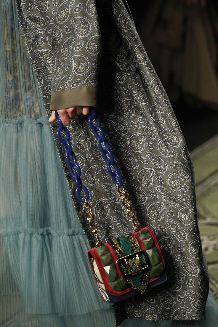 closeup shot of a model wearing a teal tulle dress layered over a longsleeved paisleyprinted coat.  the model's hand holds a small, richlydecorated handbag with a blue and gold chain strap. the handbag features a camouflage and jeweltoned design. the image focuses on the details of the clothing and accessories, showcasing the textures and patterns.  the background is blurred, suggesting a fashion show or runway setting.