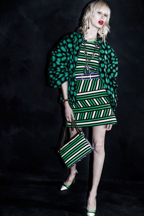 a stylish blonde model poses in a green and white striped dress paired with a matching green patterned jacket. she carries a striped handbag and wears highheeled shoes. the backdrop is dark and creates a dramatic atmosphere. the overall look is elegant and chic. the image is likely from a fashion editorial or runway show.