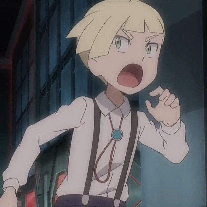 this is a close up of a young boy with blonde hair, wearing a white shirt with suspenders and a teal gem necklace. he has an open mouth, as if yelling or screaming, and a surprised expression. the background is blurry and appears to be a dark, industrial setting.