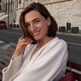 a closeup portrait of a young, attractive woman with shoulderlength brown hair. she is smiling and appears happy. she's casually dressed in a longsleeved white shirt and is sitting on a bench outdoors in a city setting.  the background features a building with architectural details. she is also carrying a small, darkred handbag. the overall mood of the image is bright and cheerful.