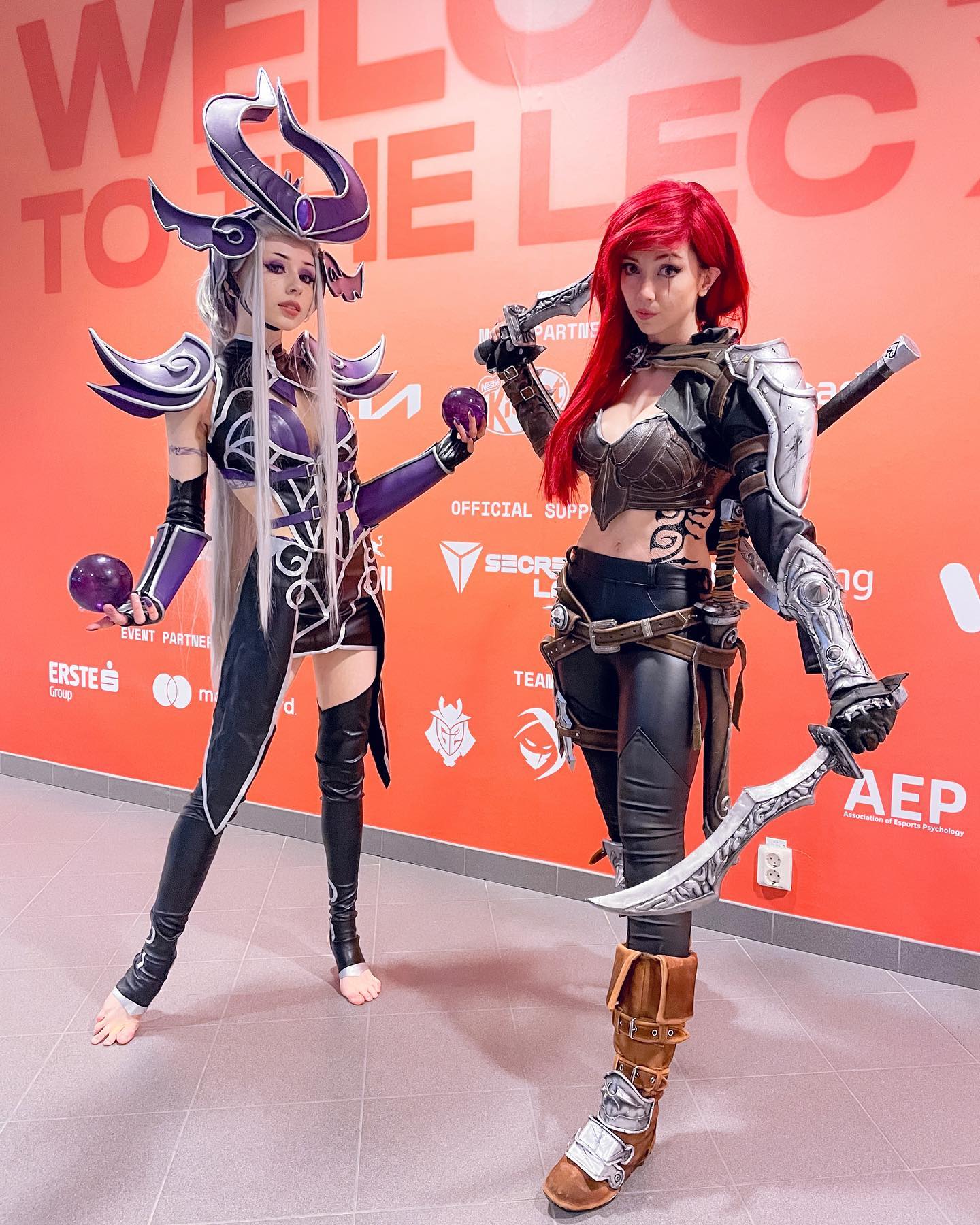 two women are cosplaying as league of legends champions, leona and riven, at an event. leona is on the left with her signature purple and white armor and long white hair. riven is on the right, in her brown and black attire with red hair. both are dressed in their characteraccurate costumes with weapons. the background has a red wall with sponsors and event logos.