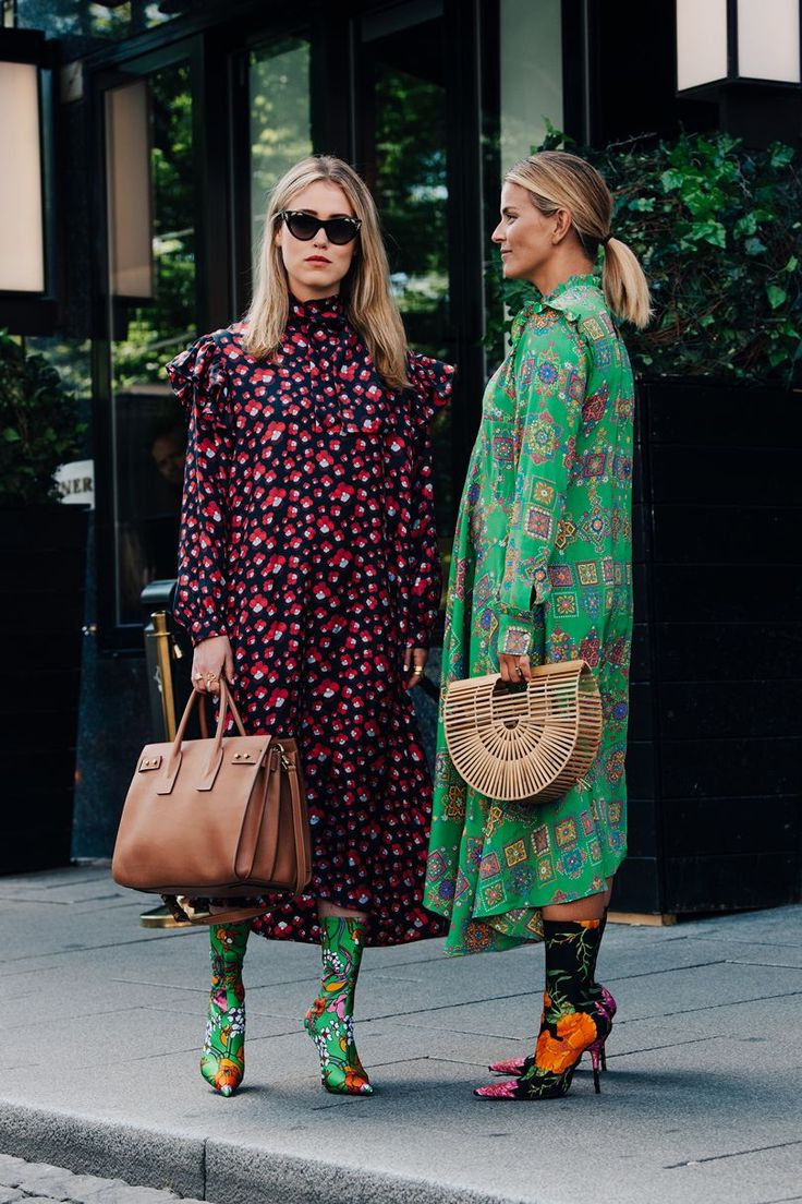 two stylish women are standing on a city sidewalk, showcasing their individual fashion sense. the woman on the left is wearing a darkcolored floral midi dress with ruffled sleeves, paired with brown leather tote bag and patterned green socks and heels. the woman on the right sports a vibrant green floral midi dress and a lightcolored woven bag, complemented with patterned socks and heels in similar color scheme.  both women are accessorized with sunglasses. they appear to be enjoying a day of shopping or a leisurely stroll. the background suggests an upscale city setting with greenery and a stylish building.