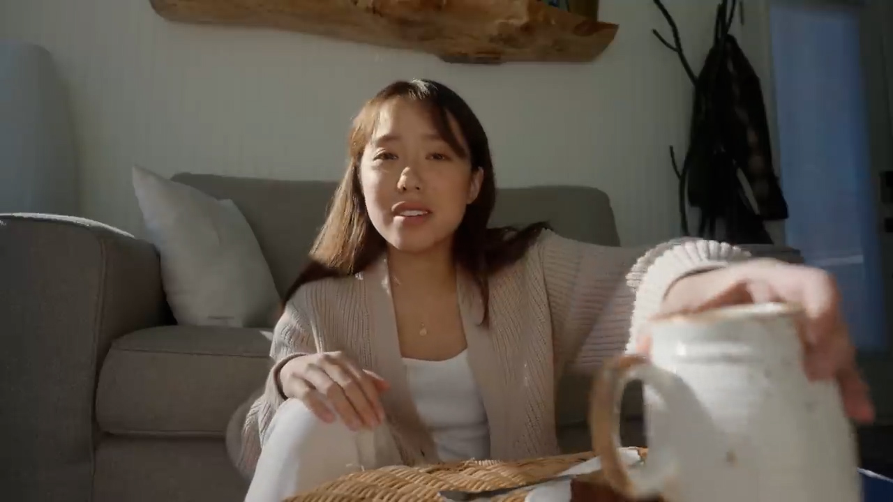 a young asian woman sits on the floor in her living room, reaching for a mug on a coffee table in front of her.  she's welllit by warm, natural light streaming in from a window behind the camera.  she's wearing a creamcolored cardigan sweater and white pants. the living room features a light grey couch with white pillows and a wooden shelf on the wall.  the overall feeling is cozy and relaxed.