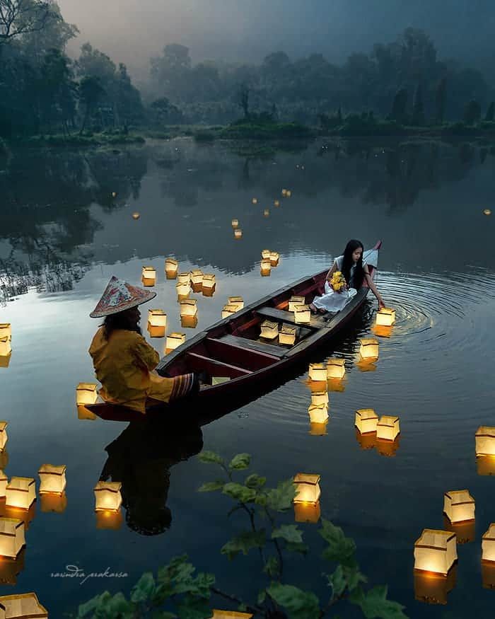 the image depicts a serene and tranquil scene at twilight or dusk on a calm lake or river. a long, narrow boat, possibly a canoe, is gently floating on the water, carrying a woman in a lightcolored dress and a man wearing a traditional asian conical hat and yellow garb.  numerous illuminated floating lanterns surround the boat, creating a magical and romantic ambiance. the surrounding landscape is peaceful and dark, with a misty or foggy atmosphere that adds to the mystical feeling. the reflection of the boat and lanterns on the water's surface enhances the scene's beauty and tranquility. the overall mood is one of peace, serenity, and perhaps a spiritual or ritualistic experience.