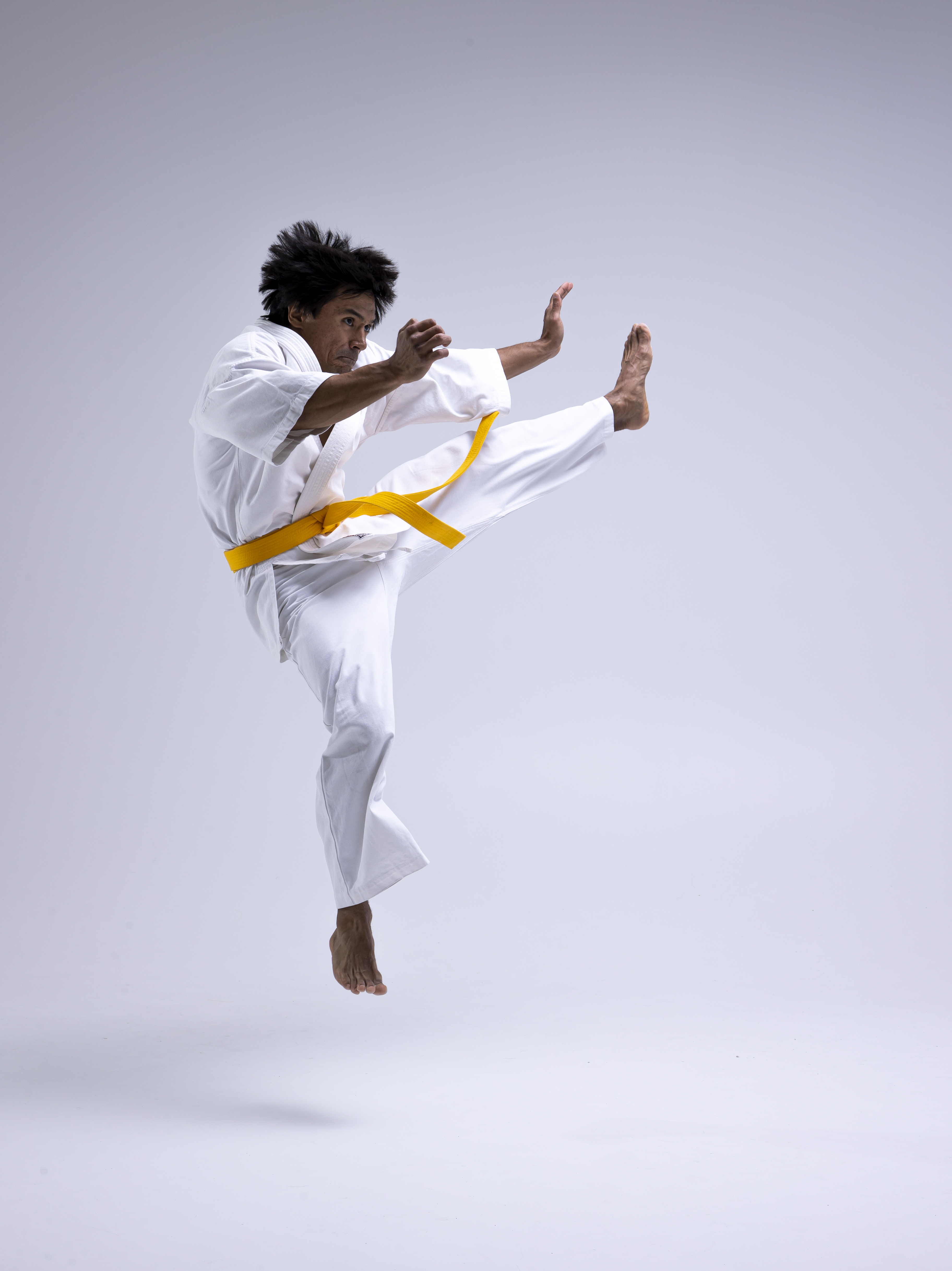 a man in a white karate uniform with a yellow belt is captured in midair, executing a high kick. he is against a light gray background.